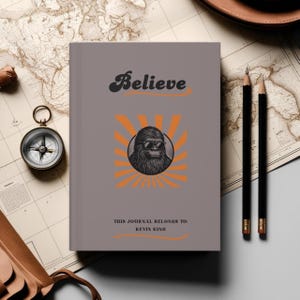 May include: A gray journal with the word "Believe" in a retro font. The cover features a cartoon Bigfoot wearing sunglasses in a circle with orange sun rays. The text "This journal belongs to: Kevin King" is printed at the bottom.