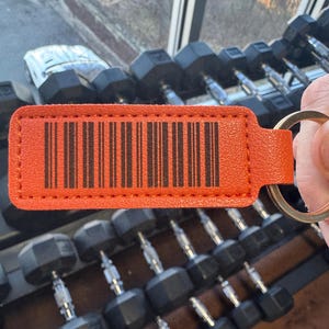 May include: Orange leather keychain with a black barcode and a silver key ring. The keychain is in focus and the background is out of focus.