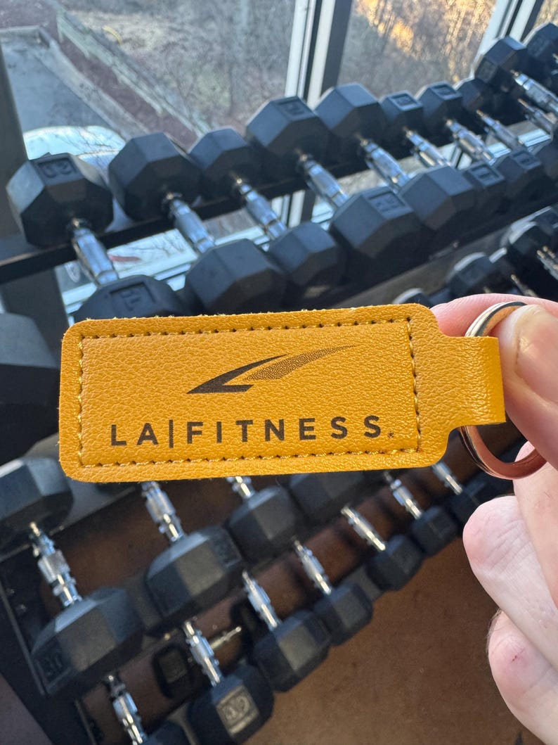 May include: A yellow leather keychain with the text "LA FITNESS" in black lettering. The keychain is hanging from a set of black dumbbells.