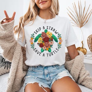 May include: White t-shirt featuring a green sea turtle surrounded by colourful flowers. The text "Skip a Straw Save a Turtle" is printed on the shirt.