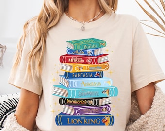 May include: A beige t-shirt with a stack of books printed on the front. The books are stacked in a pile and each book has a different Disney movie title on it. The titles are: Bambi, The Little Mermaid, Pinocchio, Fantasia, Anastasia, Snow White, Pocahontas, The Hunchback of Notre Dame, and The Lion King.