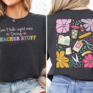 Puede incluir: Camiseta gris oscuro con el texto "Can't talk right now Doing TEACHER STUFF" en letras coloridas. La parte posterior de la camiseta presenta un diseño floral con útiles escolares, incluyendo tijeras, una regla y una taza de café.