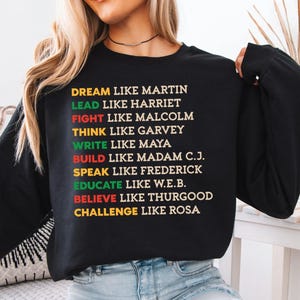 May include: Black long-sleeve sweatshirt with colorful text. The text includes phrases like "Dream like Martin," "Lead like Harriet," and "Challenge like Rosa." The text is in various colors, including yellow, green, and red.