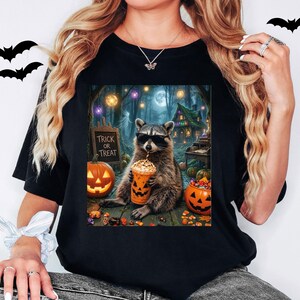 May include: A black t-shirt featuring a graphic of a raccoon wearing sunglasses and holding a pumpkin-themed cup. The scene includes a sign that reads Trick or Treat, jack-o'-lanterns, candy, and a forest background with hanging lights.