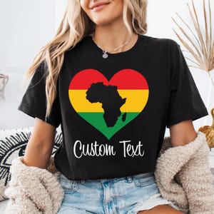 May include: Black t-shirt featuring a heart-shaped design with the African continent silhouette. The heart is colored red, yellow, and green. The words "Custom Text" are written in white script below the heart.