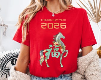 May include: Red t-shirt with gold text reading "CHINESE NEW YEAR 2026." A teal horse graphic with floral accents and Chinese characters is centered on the shirt. The shirt is a short-sleeved crew neck.