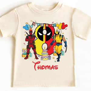 May include: A white t-shirt with a colorful graphic of Deadpool and Wolverine standing in front of a birthday cake. The number 8 is in the background and the text "Thomas" is printed below the graphic.