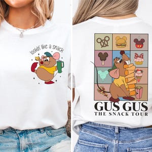 Puede incluir: Camiseta blanca con un ratón de dibujos animados sosteniendo una taza y el texto "lookin' like a snack". La parte posterior de la camiseta tiene un ratón de dibujos animados sosteniendo una pila de patatas fritas y el texto "Gus Gus The Snack Tour" con una variedad de bocadillos de dibujos animados en el fondo.