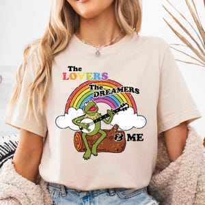 May include: A beige t-shirt with a rainbow, a green frog playing a banjo, and the text "The Lovers The Dreamers & Me".