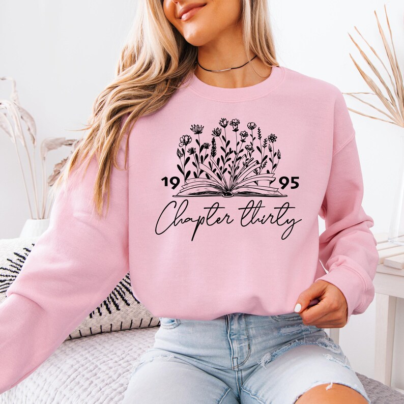 a woman sitting on a bed wearing a pink sweatshirt
