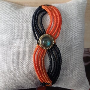 May include: A macrame bracelet featuring alternating black and orange cord strands arranged in a figure-eight pattern. A circular, metallic-rimmed cabochon stone with a dark, iridescent blue-green centre is set at the centre of the bracelet.