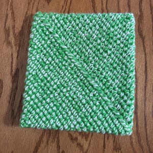 May include: A square, hand-knitted washcloth in a green and white pattern. The washcloth is made of a soft yarn and has a diagonal design element. The item is likely used for cleaning or personal hygiene.