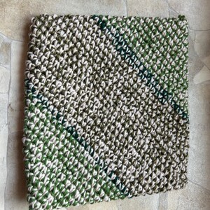 May include: Square, hand-crocheted item in shades of green, white, and olive. The textured surface features a diagonal pattern with a darker green stripe. The item is likely a decorative piece or a functional item like a trivet or coaster.