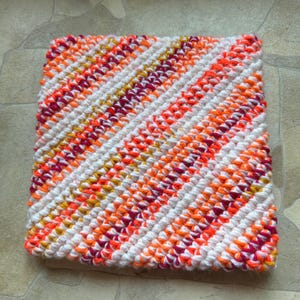 May include: A square, hand-crocheted item with a diagonal striped pattern. The design features white, orange, yellow, and burgundy yarn. The texture appears soft and the item is likely a washcloth or similar home good.
