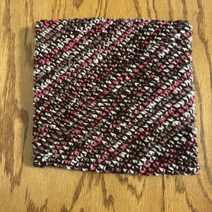 Op de afbeelding: Een vierkant, met de hand gehaakt item met een diagonaal gestreept patroon. Het kleurenschema omvat tinten bruin, wit en roze. Het item ligt op een houten oppervlak.