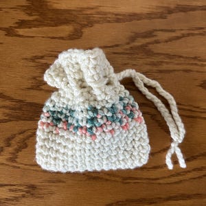 Puede incluir: Pequeño bolso de ganchillo blanco roto con cierre de cordón. El bolso presenta una banda de puntadas rosas y turquesas cerca de la parte superior. La bolsa hecha a mano probablemente se utiliza para objetos pequeños o como bolsa de regalo.