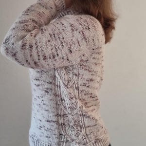 Può includere: Un maglione di lana bianco e rosa con un design a treccia sul retro. Il maglione ha un motivo maculato.