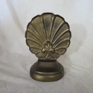 May include: A brass bookend featuring a scalloped shell design with a central fleur-de-lis motif. The object has a rounded, solid base and a metallic finish with visible vertical ridges radiating from the center to the scalloped outer edge.