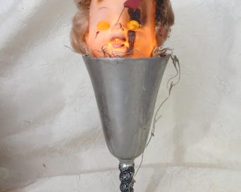 May include: A vintage doll head with light-up eyes and a red mark on its face is set inside a silver goblet. The doll has blonde hair and is a Halloween-themed decoration.