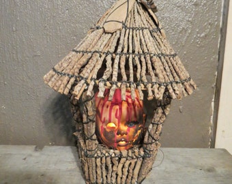 May include: A spooky Halloween decoration featuring a doll head with red paint dripping down, set inside a rustic, woven birdhouse-like structure. The doll's face is illuminated, creating a chilling effect. The structure is made of natural materials.
