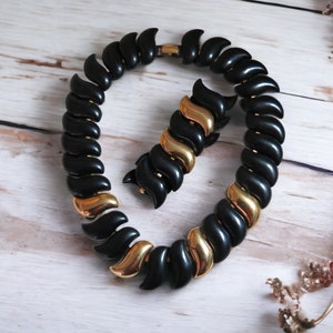 May include: A black and gold necklace and bracelet set with a swirling design. The necklace and bracelet are made of black plastic with gold accents.