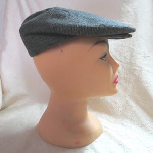 May include: A gray herringbone pattern flat cap, a type of newsboy hat, is displayed on a mannequin head. The cap has a rounded crown and a short, stiff brim. The fabric appears to be a woven wool or tweed.
