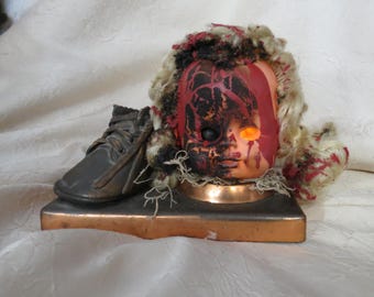 May include: A doll head with a distressed appearance, featuring red and black paint, and a single glowing orange eye. A small, dark shoe rests beside the head on a copper-colored base. The doll has blonde hair with red accents, creating a spooky aesthetic.