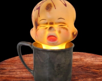 May include: A vintage-style lamp featuring a baby doll head with closed eyes and an open mouth, emitting a warm glow. The doll head is set inside a dark gray mug, creating a unique and eerie decorative piece. The background is black.