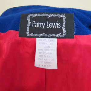 May include: A black and white label with the text "Patty Lewis" in a barbed wire frame. The label is attached to a red and blue fabric. The label also includes the text "100% Wool Flannel, Rayon Acetate Lining, Ultra Suede Trim, Dry Clean Only, Made in U.S.A., Style 421Y-STAR, Cut 7109603, Large."