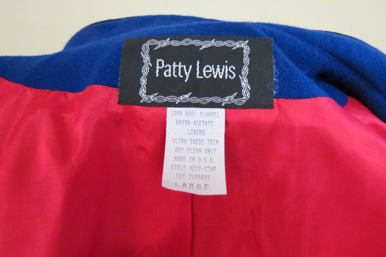 May include: A black and white label with the text "Patty Lewis" in a barbed wire frame. The label is attached to a red and blue fabric. The label also includes the text "100% Wool Flannel, Rayon Acetate Lining, Ultra Suede Trim, Dry Clean Only, Made in U.S.A., Style 421Y-STAR, Cut 7109603, Large."