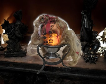 May include: A Halloween decoration featuring a doll head with blonde hair, blood-like streaks, and a glowing face. The head rests in a silver metal bowl on a decorative stand. The background includes dark decorative objects.