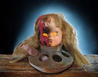 May include: A creepy doll head with blonde hair and glowing yellow eyes sits atop a rusted metal base. The doll's face is partially damaged with red and black markings, creating a spooky Halloween decoration.