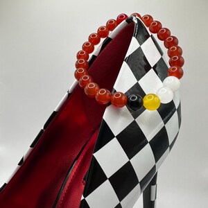 Könnte beinhalten: Ein rotes, orangefarbenes, gelbes, schwarzes und weißes Perlenarmband auf einem schwarz-weiß karierten High-Heel-Schuh.