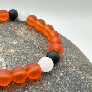 Peut inclure: Un bracelet avec des perles orange, blanches et noires. Les perles sont rondes et ont une finition mate.