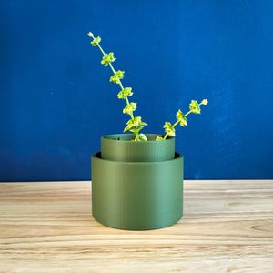 Puede incluir: Un jarrón verde oliva de dos niveles con un diseño cilíndrico. El jarrón contiene varios tallos de flores de color amarillo verdoso. El fondo es de un azul intenso. El jarrón está sobre una superficie de madera.