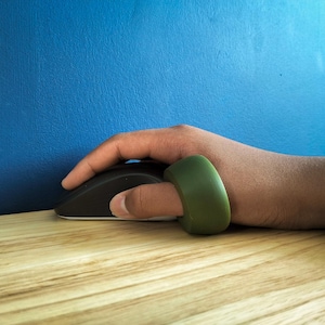 May include: A black computer mouse with a white base is held by a hand, with a green ring on the index finger. The mouse rests on a light wood surface against a blue wall.