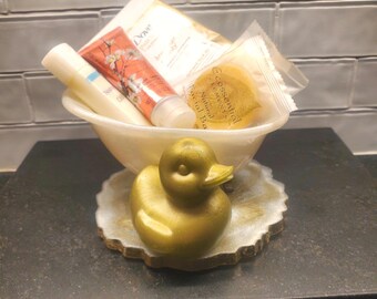 May include: A white ceramic bowl filled with bath products, including a tube of lotion, a bottle of hand cream, and a bar of soap. The bowl is sitting on a round, gray and white coaster with a gold rubber duck on top. The text on the soap reads "Ecoessential Natural Bath"
