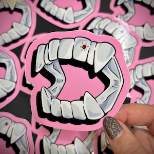 May include: A sticker featuring a set of vampire fangs with a pink background. The fangs are white with gray shading and a small red accent. Several similar stickers are visible in the background.