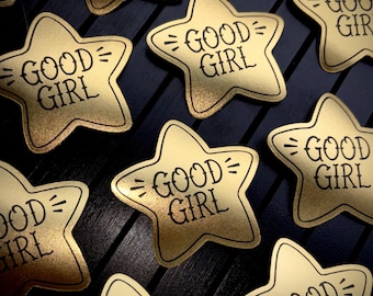 May include: A collection of star-shaped stickers with a metallic gold finish. Each sticker features the text GOOD GIRL printed in a black, stylized font, flanked by two small horizontal lines on either side of the text, centered on the star.