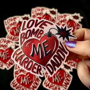 May include: A collection of stickers with a heart-shaped bomb design. The stickers are red and white, with the words "LOVE BOMB ME HARDER DADDY" in a bold font. The background is black.