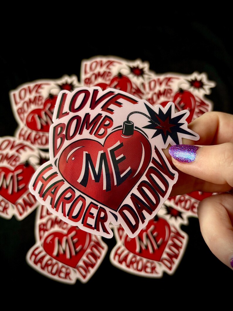 May include: A collection of stickers with a heart-shaped bomb design. The stickers are red and white, with the words "LOVE BOMB ME HARDER DADDY" in a bold font. The background is black.