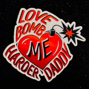 May include: A sticker with a red heart-shaped bomb design. The text reads "LOVE BOMB ME HARDER DADDY" in red and white. A black fuse and explosion graphic are also included.