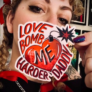 May include: A sticker with the words "LOVE BOMB ME HARDER DADDY" in red and white, featuring a heart-shaped bomb design. The sticker is held up in front of a person's face, with a silver chain necklace and red ribbons visible.