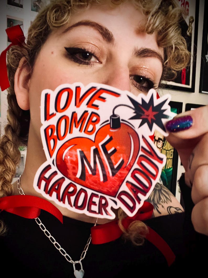 May include: A sticker with the words "LOVE BOMB ME HARDER DADDY" in red and white, featuring a heart-shaped bomb design. The sticker is held up in front of a person's face, with a silver chain necklace and red ribbons visible.