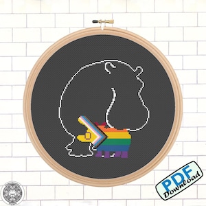 Op de afbeelding: Een zwart-wit kruissteekpatroon van een nijlpaard met een regenboogkleurig hart aan de zijkant. Het patroon is in een houten borduurring.