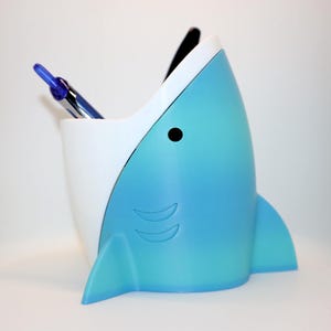 Könnte beinhalten: Ein stiftbehälter in Form eines Hais, in Blau- und Weißtönen, mit einem schwarzen Auge und Flossendetails. Ein blauer Stift ist im Inneren sichtbar. Ein lustiges und praktisches Büroaccessoire.