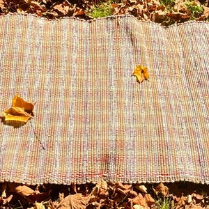 May include: A rectangular, handwoven rug with a striped pattern in shades of brown, beige, and red. Four yellow and orange autumn leaves are scattered across the rug. The rug is placed on a bed of fallen leaves.