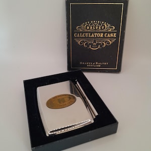 Peut inclure: Un étui à calculatrice en argent avec un logo Grant's of Dalvey en or. L'étui est dans une boîte noire avec des lettres dorées qui indiquent "The Original Grant's of Dalvey Calculator Case" et "Grants of Dalvey Scotland".