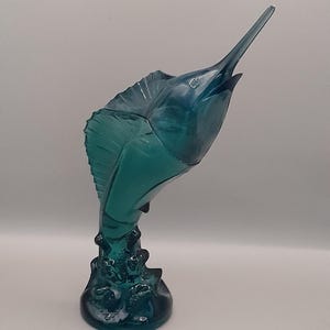 Könnte beinhalten: Eine tealblaue Glasskulptur eines Marline, der aus dem Wasser springt. Der Marlin hat einen langen, spitzen Schnabel und eine Rückenflosse. Der Sockel der Skulptur ist ein Wasserwirbel.