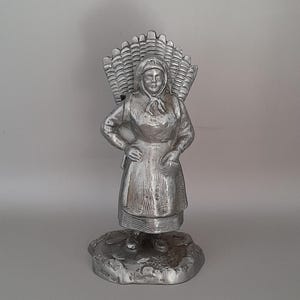 May include: A silver metal figurine of a woman wearing a long dress and a head covering, carrying a basket on her back. The figurine is standing on a textured base.
